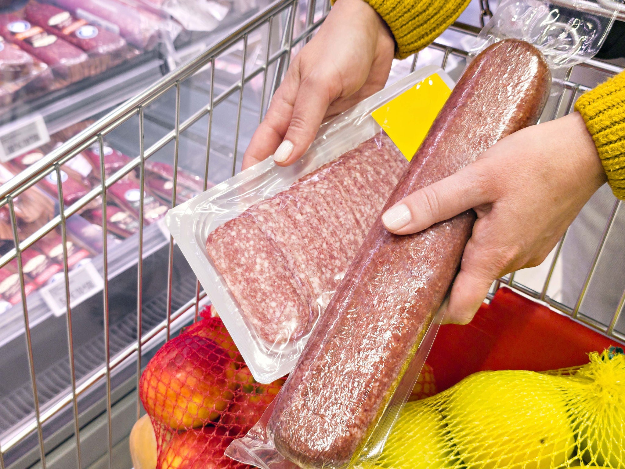 Close-up of a shopper holding packaged salami and sliced sausage over a grocery cart in a supermarket. Concept of buying deli meat, grocery shopping, and food selection; Shutterstock ID 2749477093; purchase_order: MUDE; job: ; client: Cornelia Geiger; other: ; is_generated: true