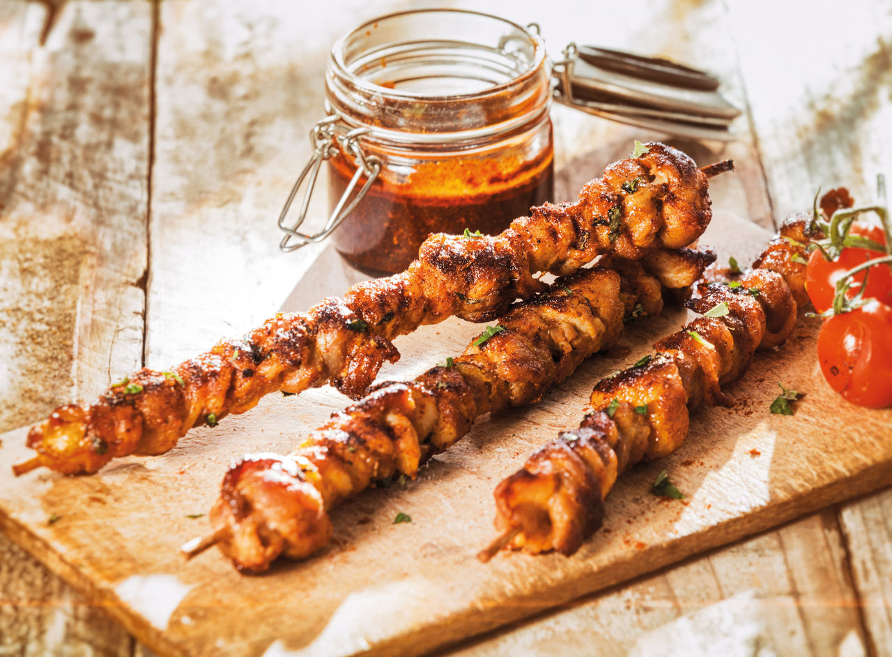 Marinated hot spicy kebabs grilled on a summer BBQ with cherry tomatoes and served on an old wooden chopping baird on a rustic weathered picnic table with a jar of basting spice